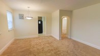 This single-floor residence at 204 D offers a straightforward and functional layout with a calm, neutral aesthetic throughout. The main living area is finished with wall-to-wall carpeting in a warm beige tone, providing a soft and cohesive foundation across the space. Walls are painted in a light, neutral shade that contributes to an airy and open feel.

A notable architectural detail is the arched doorway that connects the main living area to an adjacent room, adding a subtle classic charm to the otherwise simple design. A pendant light fixture with a clean, triangular silhouette hangs overhead, serving as a modest focal point in the dining or central area.

A doorway leads to what appears to be a kitchen, where dark cabinetry and a black refrigerator are visible, offering a contrast to the lighter tones found in the rest of the space. White baseboards run consistently throughout, providing a clean finish along the floor line.

The overall ambiance is understated and move-in ready, with a neutral palette that allows for flexible furnishing and personalization.