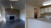 This single-floor residence offers a well-designed open-concept layout that seamlessly connects the main living areas. The living room serves as a central gathering space, anchored by a classic fireplace with a clean, white surround that adds warmth and character to the room. Dark hardwood flooring runs throughout, providing a rich contrast against the neutral, light-toned walls and lending the space a modern yet inviting feel. Vaulted or angled ceilings in the living area contribute to a sense of openness and volume, while recessed accent lighting adds a warm ambiance to the space. A peninsula-style counter with a granite or stone surface separates the living area from the kitchen, offering both a functional workspace and a casual dining or serving area. The pass-through design of the kitchen enhances the open flow of the space, allowing natural interaction between rooms. An adjacent hallway, visible through a doorway, leads to additional areas of the home. The overall aesthetic is clean and understated, with a neutral color palette, practical finishes, and thoughtful details that create a comfortable and functional living environment.