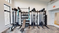 This single-floor fitness center is thoughtfully designed to support a full range of workout routines in a clean, contemporary setting. The space features soaring double-height ceilings with exposed ductwork, lending an open, industrial-modern aesthetic. Large, vertically oriented windows flood the interior with natural light and provide views of the surrounding outdoor area, creating an energizing atmosphere for exercise.

The cardio zone is positioned along the windowed wall and includes multiple treadmills and an elliptical trainer, complemented by a full-length mirror that visually expands the space. A stationary bike is situated nearby alongside built-in wooden shelving and cabinetry — complete with a beverage refrigerator — offering convenient storage and refreshment access.

The strength training area is comprehensively outfitted with cable resistance machines, a multi-tier dumbbell and kettlebell rack, medicine balls, TRX suspension straps, and an adjustable flat bench. The equipment is arranged efficiently to allow for a variety of functional and resistance-based training.

Durable stone-pattern flooring runs throughout the space, providing both practicality and visual cohesion. A set of black-framed glass double doors with transom windows offers direct access to the exterior, enhancing ventilation and connectivity to the outdoors. The overall design balances functionality with a polished, modern aesthetic.