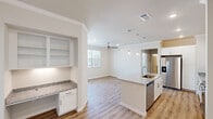 This single-story, 2-bedroom, 2-bathroom residence is designed with a clean, modern aesthetic and a functional open layout. The kitchen serves as a central highlight, featuring a large island with granite countertops, a stainless steel sink, and elegant pendant lighting overhead. White shaker-style cabinetry, a marble-tiled backsplash, and stainless steel appliances — including a refrigerator with ice maker, range, microwave, and dishwasher — complete the space. A built-in shelving nook adjacent to the kitchen adds practical storage. The kitchen flows seamlessly into the living room, which is bright and airy, benefiting from large windows with plantation shutters and a stylish ceiling fan. Light wood-tone flooring runs throughout the main living areas, contributing to a warm, cohesive feel. Both bedrooms are carpeted and include ceiling fans, generous closet space, and windows that invite natural light. The primary bedroom features a walk-in closet with wire shelving. The bathrooms are finished with granite countertops and wood-look flooring. A dedicated in-unit laundry room accommodates a full-size washer and dryer with additional wire shelving above. Crown molding accents throughout add a refined architectural touch.