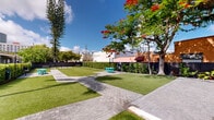 Located at 1645 NW 15th St in Miami, FL, this thoughtfully designed outdoor courtyard offers a refreshing urban retreat in the heart of South Florida. The space is defined by expansive stretches of manicured artificial turf, interspersed with geometric herringbone-patterned paver walkways that guide visitors throughout the grounds. Vibrant tropical landscaping frames the perimeter, including towering Norfolk Island pines, swaying palms, and a striking Royal Poinciana tree adorned with vivid red blooms, adding a bold splash of color against the bright Miami sky. Lush hedgerows and dark vertical fencing provide privacy and a sense of enclosure while maintaining an open, airy atmosphere. Teal-colored outdoor tables and chairs are scattered across the lawn, creating casual gathering spots ideal for socializing or relaxing. A covered pergola structure along one edge offers shaded seating with yellow chairs, providing a comfortable retreat from the sun. The space is accessed through a modern slatted metal gate, seamlessly integrating security with contemporary design. With the Miami skyline visible in the background and abundant natural light throughout the day, this courtyard presents a well-maintained, inviting outdoor environment perfectly suited to the South Florida lifestyle.