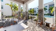 This inviting outdoor patio, located at 7130 Okeechobee Blvd in West Palm Beach, Florida, offers a well-appointed communal gathering space that seamlessly blends covered and open-air areas. The patio features a striking herringbone-patterned paver floor in a blend of charcoal and neutral tones, lending a polished and contemporary aesthetic underfoot.

A covered section with soaring ceilings is equipped with a ceiling fan, providing shade and comfort during warm Florida days. Stone-clad columns anchor the space architecturally, adding a textured, natural contrast to the crisp white modern exterior of the surrounding building. Large sliding glass doors and expansive windows along the building facade allow interior and exterior spaces to connect visually, flooding both areas with natural light.

Multiple concrete-top dining tables paired with slatted chairs accommodate generous group seating, while wooden rocking chairs positioned near the railing offer a more relaxed seating option. A gated wrought iron railing frames the perimeter of the patio, providing unobstructed views of a resort-style pool area and lush tropical landscaping filled with palm trees, ornamental shrubs, and vibrant foliage. A secured walkway with accessible ramp connects the patio to the surrounding grounds, enhancing both functionality and accessibility throughout this beautifully designed outdoor space.