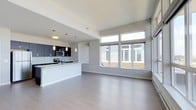 This 2-bedroom, 2-bathroom residence showcases a thoughtfully designed open-concept floor plan that seamlessly connects the kitchen and living areas into one cohesive, light-filled space. The interior is finished with smooth light-toned hardwood-style flooring that runs throughout, complementing the neutral gray and white color palette that gives the space a clean, contemporary aesthetic.

The kitchen is outfitted with dark espresso cabinetry contrasted against light countertops, creating a striking visual balance. A subway tile backsplash adds texture, while stainless steel appliances provide a polished, modern touch. Pendant lighting hangs above the kitchen peninsula, which doubles as a breakfast bar and provides additional prep space. Recessed and pendant fixtures offer layered lighting throughout.

The living area benefits from an impressive bank of oversized windows that draw in generous amounts of natural light and offer outward views to the surroundings. The elevated window placement ensures both brightness and privacy. Elevated ceilings further enhance the sense of openness and volume in the main living space. The overall ambiance is modern, airy, and functional, designed for comfortable everyday living.