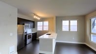This single-floor space showcases a contemporary open-concept layout that seamlessly integrates the kitchen and living area. The kitchen is appointed with dark espresso cabinetry, a subway tile backsplash, and a full suite of stainless steel appliances, including a refrigerator, electric range, and built-in microwave. A sleek white waterfall island anchors the space, offering an undermount sink with a modern arc faucet and generous counter space for meal preparation or casual dining. Track lighting overhead provides focused illumination throughout the kitchen zone. Dark hardwood flooring runs throughout, creating a striking contrast against the light-toned walls and white island surface. Multiple windows allow an abundance of natural light to fill the space while framing urban cityscape views, suggesting an elevated floor position within a multi-story building. The open living area adjacent to the kitchen benefits from this same natural light, contributing to an airy, connected atmosphere. Overall, the space presents a clean, modern aesthetic well-suited for urban living.