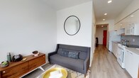 This single-floor space showcases a thoughtfully designed interior that blends modern minimalism with mid-century modern touches. The living area serves as the heart of the space, featuring light wood-toned flooring that runs throughout, creating a warm and cohesive feel underfoot. Crisp white walls provide a clean backdrop, enhanced by recessed ceiling lighting that keeps the atmosphere bright and airy.

A charming seating area anchors the living space, highlighted by a contemporary gray sofa accented with a textured throw pillow. A circular black-framed mirror mounted above the sofa adds a stylish focal point, visually expanding the room. A warm-toned wood media console sits alongside, lending a mid-century character to the space, while a gold accent tray table adds a touch of modern sophistication.

A hallway extends from the living area, leading further into the unit with continued light flooring and recessed lighting overhead. A bold red door at the end of the corridor provides a striking pop of color, serving as a distinctive architectural accent that adds personality to the otherwise neutral palette.

Overall, this space offers a well-curated, functional layout with a clean aesthetic that balances comfort and contemporary style seamlessly.