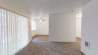 This single-floor space offers a straightforward, functional layout with clearly defined living areas. The main living area features wall-to-wall carpeting underfoot and is bathed in natural light streaming through a large window dressed with vertical blinds, contributing to a bright and open atmosphere. A textured popcorn ceiling adds a classic touch overhead throughout the space. An open passageway connects the carpeted living area to an adjacent room finished with hardwood-style flooring, creating a natural visual distinction between the two spaces. This secondary area is equipped with a ceiling fan fitted with a light fixture, providing both air circulation and overhead lighting. The walls throughout are painted in a clean, neutral white, offering a versatile backdrop suitable for a variety of interior styles. Baseboard trim runs along the base of the walls, adding a finished look to the rooms. The space also includes a smoke detector, reflecting attention to basic safety standards. Overall, the unit presents a clean, move-in-ready environment with practical features and a simple, uncluttered design aesthetic throughout its single level.