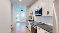 This well-appointed single-floor unit offers a thoughtfully designed layout with modern finishes throughout. The galley-style kitchen is a standout feature, showcasing crisp white shaker cabinetry, dark granite countertops, a herringbone-patterned tile backsplash, and a full suite of stainless steel appliances including a range, microwave, dishwasher, and refrigerator. A matte black faucet adds a refined contrast to the sink area. Warm-toned hardwood-style flooring runs continuously throughout the space, lending a cohesive and inviting feel. The open corridor leading from the kitchen connects seamlessly to the main living area, which is brightened by large windows dressed with blinds and features a ceiling fan overhead. A dedicated laundry area includes a stacked washer and dryer alongside built-in shelving, providing convenient storage. The bathroom mirrors the kitchen's design language with matching white cabinetry, granite countertops, an undermount sink, and matte black fixtures, complemented by a tall linen cabinet for additional storage. Recessed lighting and clean white walls enhance the bright, contemporary ambiance throughout the entire unit.