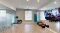 This dedicated yoga and fitness room offers a clean, open layout designed to support a wide range of exercise and wellness activities. The space features smooth hardwood-style flooring that provides an ideal surface for yoga, stretching, and functional training. Neutral gray walls and white trim create a calm, focused atmosphere, while recessed lighting throughout the ceiling ensures bright, even illumination during any time of day.

Natural light filters in through multiple windows fitted with white blinds, adding an airy quality to the room. A large wall-to-wall mirror spans one side of the space, enhancing the sense of openness and allowing practitioners to monitor their form during workouts. Two wall-mounted flat-screen TVs are positioned for easy viewing from the floor, supporting guided yoga sessions, virtual fitness classes, or instructional content.

The room comes well-stocked with a variety of fitness equipment, including a tiered rack of colorful kettlebells in multiple weights, several stability and medicine balls, and a rolled yoga mat for individual use. A small speaker system is also present, allowing for an immersive audio experience during sessions. The overall ambiance is welcoming and purposeful, making this an excellent space for yoga, strength training, stretching, or mindful movement.
