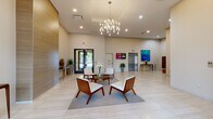 This contemporary building lobby makes a polished first impression with its expansive, open layout and soaring ceilings that lend the space a grand, airy quality. Large-format light stone tile flooring runs throughout, reflecting the abundant recessed lighting and contributing to the bright, clean aesthetic. At the heart of the lobby, a statement chandelier with candelabra-style arms serves as a striking focal point, anchoring a central seating arrangement composed of mid-century modern armchairs in white upholstery with warm wood frames, gathered around a circular glass-top coffee table atop a soft area rug.

The walls feature carefully curated large-scale artwork, including vibrant abstract canvases in blue and warm tones, adding color and visual interest to the neutral palette. A textured wood-panel accent wall introduces warmth and architectural depth. Lush potted plants are positioned throughout, bringing a touch of greenery to the refined interior.

Practical amenities are seamlessly integrated: a stainless steel elevator provides vertical access, a bank of resident mailboxes is neatly organized along one wall, and a digital display screen offers building information. Floor-to-ceiling glass entry doors with black metal frames allow natural light to filter in while maintaining views to the outdoors, completing this welcoming and functional lobby environment.