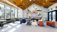 This thoughtfully designed community lounge offers a spacious, open-plan interior that makes a striking first impression. The architecture centers on a dramatic vaulted ceiling clad in warm natural wood planks, supported by exposed beams and fitted with recessed track lighting — a standout feature that draws the eye upward and amplifies the sense of volume throughout the room.

Large floor-to-ceiling windows and glass-panel folding doors line the exterior walls, flooding the interior with abundant natural light and providing seamless visual and physical access to an outdoor pool and patio area beyond. Clerestory windows above the doors further enhance the brightness of the space.

The interior is organized into several distinct seating zones, each thoughtfully furnished with a vibrant, eclectic mix of chairs, sofas, and accent pieces in bold navy, terracotta, yellow, and grey tones. A focal media wall features a large flat-screen display framed by floating wood shelves adorned with curated artwork and decorative objects, set against a patterned accent wall.

Additional zones include a casual bar-height dining area near the windows, a booth-style banquette seating nook, and open lounge areas ideal for socializing or relaxing. Dark green tiled accent walls with open shelving provide functional and aesthetic contrast throughout. The polished concrete and large-format tile flooring ties the entire space together with a clean, modern finish.