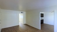This single-story space presents a clean, minimalist layout designed for functional everyday living. The main living area features warm wood-style laminate flooring that extends throughout, complemented by crisp white walls and a neutral cream ceiling that together create a bright, airy atmosphere. An HVAC vent is integrated into the ceiling, ensuring climate comfort year-round. The open layout connects seamlessly to adjacent spaces, with a wide doorway leading to a hallway that provides access to additional rooms. To the right, an open passage reveals a glimpse of the kitchen, which is outfitted with dark-toned cabinetry and light countertops, offering a notable contrast to the otherwise light color palette. The kitchen's design suggests practical storage and workspace. The overall ambiance of the space is understated and move-in ready, with a straightforward floor plan that allows for flexible furniture arrangement. The consistent flooring and neutral tones throughout contribute to a cohesive visual flow from room to room, making the space feel unified and well-maintained.
