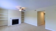 This single-floor space centers around a spacious living area finished with wall-to-wall carpeting that adds warmth underfoot. The room is anchored by a white-mantled fireplace with a sleek black surround, providing a classic focal point along one wall. Adjacent to the fireplace, a set of built-in open shelves offers convenient display and storage options, seamlessly integrated into the wall for a clean, cohesive look. A ceiling fan with an integrated light fixture is centrally mounted, promoting air circulation and supplemental lighting throughout the room. The walls are painted in a neutral beige tone, complementing the light carpeting and contributing to a bright, airy atmosphere. A textured popcorn ceiling adds a traditional touch overhead. An open passageway leads to an adjoining room or hallway, suggesting a practical, connected floor plan. Visible HVAC vents indicate a climate-controlled environment. White baseboards frame the perimeter of the room, providing a crisp contrast to the neutral walls and flooring. Overall, the space offers a straightforward, move-in-ready layout suited to comfortable everyday living.