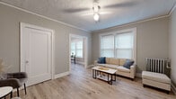This single-floor space is thoughtfully laid out across several connected rooms, each finished with light hardwood-style flooring and soft, neutral gray and beige wall tones that create a cohesive and calming atmosphere throughout. Crown molding and crisp white baseboards add a clean, classic detail to the interior.

The living room serves as the central gathering space, featuring two windows fitted with white horizontal blinds that allow ample natural light to filter in. A ceiling fan with a light fixture provides both comfort and illumination. The textured ceiling adds a traditional architectural touch consistent throughout the unit. An open doorway connects the living room directly to the dining area, which is bright and windowed with a double-window arrangement, offering a pleasant outdoor view.

The kitchen is compact and functional, equipped with granite-style countertops, white cabinetry, and a stainless steel sink. The bedroom is a well-proportioned, quiet retreat with a window providing natural light and continued hardwood-style flooring. The bathroom includes a tiled tub-shower combination, a window for ventilation, and a classic radiator heater, adding a charming vintage character to the space.