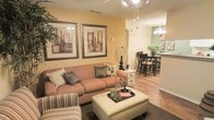 This single-floor residence offers an inviting, open-concept layout that seamlessly connects the main living, dining, and kitchen areas. The living room is warm and well-appointed, featuring rich hardwood-style flooring, a textured ceiling, and walls painted in a soft golden hue that lends the space a welcoming ambiance. A plush, earth-toned sofa paired with coordinating throw pillows anchors the seating area, complemented by a large upholstered ottoman that doubles as a central coffee table. Framed botanical artwork adorns the walls, adding a decorative focal point. Track lighting overhead provides functional illumination throughout the open space. Flowing naturally from the living area, the dining space accommodates a dark-wood dining table and chairs, ideal for everyday meals or entertaining. A decorative chandelier with a classic multi-bulb design hangs above, enhancing the room's charm. Just beyond, the kitchen features a raised breakfast bar, sage-green accent walls, and pendant lighting, creating a distinct yet connected culinary space. Lush greenery in the form of a potted plant adds a fresh, natural touch to the dining and kitchen area. The overall design balances comfort and style with a cohesive, neutral color palette throughout.
