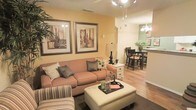 This single-floor residence offers an inviting, open-concept layout that seamlessly connects the main living, dining, and kitchen areas. The living room is warm and well-appointed, featuring rich hardwood-style flooring, a textured ceiling, and walls painted in a soft golden hue that lends the space a welcoming ambiance. A plush, earth-toned sofa paired with coordinating throw pillows anchors the seating area, complemented by a large upholstered ottoman that doubles as a central coffee table. Framed botanical artwork adorns the walls, adding a decorative focal point. Track lighting overhead provides functional illumination throughout the open space. Flowing naturally from the living area, the dining space accommodates a dark-wood dining table and chairs, ideal for everyday meals or entertaining. A decorative chandelier with a classic multi-bulb design hangs above, enhancing the room's charm. Just beyond, the kitchen features a raised breakfast bar, sage-green accent walls, and pendant lighting, creating a distinct yet connected culinary space. Lush greenery in the form of a potted plant adds a fresh, natural touch to the dining and kitchen area. The overall design balances comfort and style with a cohesive, neutral color palette throughout.