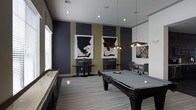 This single-floor game room offers a well-appointed recreational space designed with a contemporary aesthetic. The room's most prominent feature is a full-sized pool table anchored at the center, illuminated by a striking multi-bulb pendant light fixture that adds both function and visual interest. To one side, a shuffleboard table runs along the wall, providing an additional entertainment option for guests.

The interior design is defined by bold, dark charcoal accent walls contrasted with warm olive-gold vertical stripes, creating a dynamic and sophisticated atmosphere. Two framed abstract artworks are symmetrically displayed on the accent wall, reinforcing the modern design sensibility. A third piece of wall art adds further visual depth to the space.

Recessed ceiling lighting, complemented by the pendant fixture, ensures the room is well-lit throughout. High-top bistro tables with bar stools are positioned along the walls, offering casual seating for spectators or social gatherings. A pair of upholstered lounge chairs provide a more relaxed seating option. Tile flooring bordered by a patterned area rug ties the design elements together cohesively.