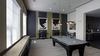 This single-floor game room offers a well-appointed recreational space designed with a contemporary aesthetic. The room's most prominent feature is a full-sized pool table anchored at the center, illuminated by a striking multi-bulb pendant light fixture that adds both function and visual interest. To one side, a shuffleboard table runs along the wall, providing an additional entertainment option for guests.

The interior design is defined by bold, dark charcoal accent walls contrasted with warm olive-gold vertical stripes, creating a dynamic and sophisticated atmosphere. Two framed abstract artworks are symmetrically displayed on the accent wall, reinforcing the modern design sensibility. A third piece of wall art adds further visual depth to the space.

Recessed ceiling lighting, complemented by the pendant fixture, ensures the room is well-lit throughout. High-top bistro tables with bar stools are positioned along the walls, offering casual seating for spectators or social gatherings. A pair of upholstered lounge chairs provide a more relaxed seating option. Tile flooring bordered by a patterned area rug ties the design elements together cohesively.