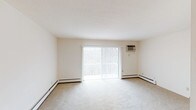 This single-floor unit offers a clean, straightforward layout spread across several well-defined rooms. The living area is generously sized, featuring neutral wall-to-wall carpeting, baseboard heating, and a wall-mounted air conditioning unit. A large sliding glass door floods the space with natural light and provides access to an outdoor area, with views of surrounding trees visible through the glass.

The kitchen is practical and well-equipped, featuring warm oak cabinetry extending to the upper and lower walls, light granite-style countertops offering ample prep space, hardwood flooring, and a coil-top electric range. The layout creates an efficient work triangle with good counter space throughout.

The bedrooms are carpeted and finished in neutral tones, each equipped with ceiling fan light fixtures. One bedroom includes a walk-in closet with oak shelving, while others feature standard closet configurations. Baseboard heating units and natural-light windows are present throughout the sleeping areas.

The bathroom includes a full soaking tub with shower, accented by blue mosaic tile on the walls, white fixtures, and a large vanity mirror. Overall, the unit maintains a clean, functional aesthetic with consistent neutral finishes throughout.