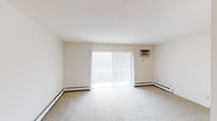 This single-floor unit offers a clean, straightforward layout spread across several well-defined rooms. The living area is generously sized, featuring neutral wall-to-wall carpeting, baseboard heating, and a wall-mounted air conditioning unit. A large sliding glass door floods the space with natural light and provides access to an outdoor area, with views of surrounding trees visible through the glass.

The kitchen is practical and well-equipped, featuring warm oak cabinetry extending to the upper and lower walls, light granite-style countertops offering ample prep space, hardwood flooring, and a coil-top electric range. The layout creates an efficient work triangle with good counter space throughout.

The bedrooms are carpeted and finished in neutral tones, each equipped with ceiling fan light fixtures. One bedroom includes a walk-in closet with oak shelving, while others feature standard closet configurations. Baseboard heating units and natural-light windows are present throughout the sleeping areas.

The bathroom includes a full soaking tub with shower, accented by blue mosaic tile on the walls, white fixtures, and a large vanity mirror. Overall, the unit maintains a clean, functional aesthetic with consistent neutral finishes throughout.