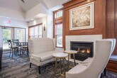 This sophisticated single-floor community space offers a generously sized, multi-functional layout designed for both socializing and private gatherings. The main lounge area serves as the heart of the space, featuring soaring tray ceilings adorned with elegant chandelier lighting, plush gray sofas, and an entertainment console with a wall-mounted flat-screen TV. Patterned carpeting in muted blue and gray tones flows throughout, tying the interior together cohesively.

Multiple seating and dining clusters are thoughtfully arranged across the open floor plan, including round tables with upholstered chairs suited for gaming or meetings. Rich warm wood paneling, a fireplace nook with wingback chairs, and framed artwork lend a refined, club-like ambiance. Wooden-trimmed windows with slatted blinds line the walls, inviting generous natural light and views of the surrounding greenery.

Two semi-private rooms offer additional space for smaller group gatherings or meetings, each with its own round table seating and direct access to an outdoor patio via glass doors. The fully equipped kitchen features stainless steel appliances, white cabinetry, dark countertops, and a peninsula with bar seating — ideal for catering or casual use. Together, these areas create a versatile and elegantly designed community environment.