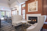 This sophisticated single-floor community space offers a generously sized, multi-functional layout designed for both socializing and private gatherings. The main lounge area serves as the heart of the space, featuring soaring tray ceilings adorned with elegant chandelier lighting, plush gray sofas, and an entertainment console with a wall-mounted flat-screen TV. Patterned carpeting in muted blue and gray tones flows throughout, tying the interior together cohesively.

Multiple seating and dining clusters are thoughtfully arranged across the open floor plan, including round tables with upholstered chairs suited for gaming or meetings. Rich warm wood paneling, a fireplace nook with wingback chairs, and framed artwork lend a refined, club-like ambiance. Wooden-trimmed windows with slatted blinds line the walls, inviting generous natural light and views of the surrounding greenery.

Two semi-private rooms offer additional space for smaller group gatherings or meetings, each with its own round table seating and direct access to an outdoor patio via glass doors. The fully equipped kitchen features stainless steel appliances, white cabinetry, dark countertops, and a peninsula with bar seating — ideal for catering or casual use. Together, these areas create a versatile and elegantly designed community environment.