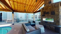 This well-appointed outdoor amenity space combines comfort and functionality in a resort-inspired setting. At its heart is a generously sized covered pavilion with a striking vaulted wood-beam ceiling crafted from warm-toned timber, lending the area a lodge-like ambiance. Beneath the pavilion, a large stone fireplace serves as a dramatic focal point, flanked by wicker lounge seating with plush cushions and natural wood stump side tables, creating an inviting gathering space for residents. A gas BBQ grill is conveniently positioned nearby, making outdoor cooking effortless.

The pavilion opens directly onto an expansive concrete pool deck, where a sparkling outdoor swimming pool takes center stage. The pool is enclosed by a safety fence and surrounded by lounge chairs for sunbathing under open skies. Adjacent to the covered area, a bubbling hot tub provides an additional relaxation option. A secondary pergola structure is visible across the deck, offering additional shaded seating.

Stone accents on the support columns and fireplace echo throughout the design, tying together the natural materials palette. Lush tropical plantings, including palm trees, add greenery and a resort-like feel to the surroundings. The space is thoughtfully designed to encourage community gathering and outdoor leisure throughout the seasons.