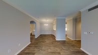 This single-floor residence showcases a thoughtfully designed interior that balances classic elegance with functional living. Rich hardwood-style flooring runs throughout the main living areas, providing a warm and cohesive foundation for the space. Crown molding adorns the ceilings, adding a refined architectural detail that elevates the overall aesthetic. A defining feature of the dining area is the decorative wainscoting along the lower walls, complemented by a stylish chandelier that serves as a focal point overhead. Structural columns and a graceful arched doorway introduce subtle architectural character, creating natural transitions between spaces without sacrificing the open feel of the floor plan. The neutral, light gray wall palette keeps the interior feeling bright and airy, allowing the warm tones of the flooring to stand out. A glimpse through the arched entryway reveals an adjacent room with white cabinetry, suggesting a well-appointed utility or bathroom space nearby. The open layout promotes a seamless flow between the living, dining, and kitchen areas, making the home feel spacious and inviting. Overall, the interior presents a clean, move-in-ready environment with timeless design touches throughout.