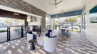Located in Ocala, Florida, this single-level covered outdoor amenity space offers a well-appointed environment designed for relaxation and social gatherings. The space features a distinctive patterned brick paver flooring that runs throughout, lending a refined yet durable foundation to the area.

The lounge zone is anchored by contemporary curved lounge chairs in white and navy blue, arranged around a cylindrical concrete coffee table, providing a stylish and comfortable seating arrangement. Complementing this is a dedicated dining area outfitted with a long rectangular table and matching chairs, ideal for group gatherings. Ceiling fans are mounted throughout the covered portions of the space, ensuring airflow and comfort in the Florida climate.

A striking stacked-stone accent wall serves as a prominent architectural feature, adding texture and visual contrast against the cool blue-gray painted walls. Large-format tile cladding on an adjacent wall adds a modern touch, and a wall-mounted flat-screen television enhances the space's functionality as an entertainment area. Black metal railing encloses the perimeter, offering safety while preserving open sightlines to lush palm trees, green landscaping, and the surrounding residential community. Glass doors connect the space to an interior clubhouse or common area, making it a seamlessly integrated extension of the broader amenity offerings.