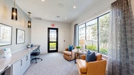 Located in Houston, TX (77407), this single-floor living room serves as a versatile lounge and workspace, thoughtfully designed to balance comfort and productivity. The room is bright and welcoming, with crisp white walls, recessed lighting, and plush patterned carpeting that adds warmth underfoot.

A standout feature is the expansive floor-to-ceiling window wall, framed by sheer drape panels, which floods the space with natural light and offers clear views of an outdoor pool area and surrounding landscaping. A glass-paneled exterior door further enhances the connection to the outdoors.

The seating area is anchored by tan leather armchairs accented with bold blue and teal throw pillows, arranged around a round side table with a classic table lamp and a small potted plant — creating an inviting spot for relaxation or casual conversation.

Along the opposite wall, a long built-in desk with lower cabinetry provides ample workspace for two, complemented by ergonomic task chairs and pendant lighting in a deep, smoky finish. Framed artwork depicting colorful scenes adds a decorative touch above the workstation.

The overall ambiance is modern and polished, combining functional design elements with carefully chosen furnishings to create a space that is both practical and aesthetically appealing.