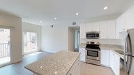 This single-floor residence offers a clean, contemporary design centered around an open-concept layout that seamlessly connects the kitchen and living areas. The kitchen is a standout feature, showcasing crisp white cabinetry that extends to the ceiling, light-toned granite countertops with subtle veining, and a central island that provides generous prep space and casual seating. Stainless steel appliances, including a range with an electric cooktop and an over-the-range microwave, lend a polished, modern finish to the space. Recessed lighting is thoughtfully positioned throughout the ceiling, complementing the abundant natural light that streams in through the windows. The light hardwood flooring runs continuously through the kitchen and into the adjoining living area, reinforcing the open, airy feel of the floor plan. Neutral white walls and smooth ceilings maintain a bright, uncluttered atmosphere throughout. A hallway visible from the main living area hints at additional rooms beyond, suggesting a well-organized and functional layout. The overall ambiance is fresh, modern, and move-in ready, making this space well-suited for everyday living and entertaining alike.