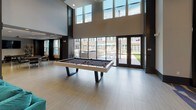 Located at 15250 Siesta Key Way in Rockville, Maryland, this single-floor amenity space delivers a bold, contemporary aesthetic defined by high ceilings, dramatic finishes, and thoughtfully curated furnishings throughout. The entry corridor immediately sets the tone with a striking floor-to-ceiling marble accent wall featuring sweeping natural veining, flanked by statement hood chairs and a patterned carpet that transitions elegantly to large-format light tile flooring. The main lounge area commands attention with soaring double-height ceilings, an oversized circular pendant light fixture, a wall-mounted flat-screen TV, and a sleek linear fireplace — all anchored by a generous teal sectional sofa. Adjacent to this is a sophisticated entertainment nook showcasing a sculptural chandelier, a silver elephant wall sculpture, curated artwork, and plush black swivel chairs arranged around a metallic ottoman. A separate lounge zone features a coffered ceiling with recessed lighting, neutral upholstered seating, mirrored accent tables, and tall decorative planters that add organic warmth. The game area is bright and open, highlighted by floor-to-ceiling glass doors and clerestory windows that flood the space with natural light and offer direct views and access to an outdoor courtyard, where a modern billiards table serves as a central recreational feature.
