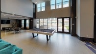 Located at 15250 Siesta Key Way in Rockville, Maryland, this single-floor amenity space delivers a bold, contemporary aesthetic defined by high ceilings, dramatic finishes, and thoughtfully curated furnishings throughout. The entry corridor immediately sets the tone with a striking floor-to-ceiling marble accent wall featuring sweeping natural veining, flanked by statement hood chairs and a patterned carpet that transitions elegantly to large-format light tile flooring. The main lounge area commands attention with soaring double-height ceilings, an oversized circular pendant light fixture, a wall-mounted flat-screen TV, and a sleek linear fireplace — all anchored by a generous teal sectional sofa. Adjacent to this is a sophisticated entertainment nook showcasing a sculptural chandelier, a silver elephant wall sculpture, curated artwork, and plush black swivel chairs arranged around a metallic ottoman. A separate lounge zone features a coffered ceiling with recessed lighting, neutral upholstered seating, mirrored accent tables, and tall decorative planters that add organic warmth. The game area is bright and open, highlighted by floor-to-ceiling glass doors and clerestory windows that flood the space with natural light and offer direct views and access to an outdoor courtyard, where a modern billiards table serves as a central recreational feature.