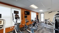 This fitness center is designed to provide a functional and motivating workout environment. The space features bold burnt-orange accent walls that energize the room, contrasting with neutral gray tones on the ceiling and remaining walls. A drop-panel ceiling with recessed fluorescent lighting ensures bright, even illumination throughout. The flooring consists of durable, patterned carpet tiles suited for high-traffic fitness use. Large windows along the walls allow natural light to filter in, creating an open and welcoming atmosphere. The cardio equipment lineup includes elliptical trainers, upright and recumbent stationary bikes, and additional exercise machines, all arranged for easy access and comfortable use. Flat-screen televisions are mounted on the accent wall — including one large display and a smaller monitor — allowing users to stay entertained during workouts. A glass-paneled door provides direct access to an exterior area, adding a sense of connectivity to the outdoors. The overall layout is efficient and thoughtfully organized, making the most of the available space while maintaining clear pathways between equipment. This fitness center offers a clean, modern setting suitable for a variety of cardio-focused exercise routines.