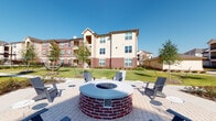Located at 5330 Sienna Pkwy in Missouri City, Texas, this thoughtfully designed outdoor amenity space serves as a central gathering hub within a well-appointed residential community. The space is defined by its classic brick construction, which carries consistently throughout the exterior walls, columns, and accent details, lending a cohesive and polished aesthetic.

At the heart of the outdoor area is a prominent circular fire pit built with a brick base and filled with decorative blue glass, surrounded by Adirondack-style chairs arranged on a paved stone patio — ideal for relaxed social gatherings. Nearby, a covered outdoor lounge area features brick columns supporting an overhead structure, with a wall-mounted television, comfortable seating, and an iron railing that frames views of the manicured courtyard and multi-story residential buildings beyond.

A paved brick walkway corridor connects various points of the space, flanked by floor-to-ceiling glass sliding doors that allow natural light to flow between interior and exterior areas. Accent plantings, including snake plants and other greenery, add life to the transitional spaces. A secured gated entry with ornamental iron fencing provides both safety and architectural character.

Throughout, the space balances functional outdoor living with refined design details, offering residents inviting areas for relaxation and community engagement under open Texas skies.
