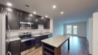 This thoughtfully designed single-floor residence offers a seamless blend of modern finishes and functional living spaces. The kitchen serves as the heart of the home, showcasing rich dark cabinetry, light granite countertops, a full suite of stainless steel appliances including a refrigerator, range, microwave, and dishwasher, and a generously sized granite-topped island with built-in shelving. Recessed lighting illuminates the workspace, while wood-look flooring ties the kitchen into the adjacent living area. The open living space is bright and airy, benefiting from natural light that pours in through French doors, which lead to an outdoor balcony and provide views of the surrounding landscape. A convenient built-in closet with wire shelving offers ample storage just off the living area. The bedroom is a comfortable retreat featuring plush carpeting, two windows with outdoor views, and a clean, neutral palette. The bathroom is finished with a classic black-and-white mosaic tile floor, a dark vanity with a white countertop, a large mirror, and a shower-tub combination. A dedicated laundry area with a stacked washer and dryer unit, complemented by additional wire shelving, adds everyday convenience to this well-appointed residence.