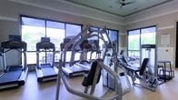 This fitness center is designed to support a comprehensive workout experience within a bright, open layout. The space is outfitted with multiple treadmills positioned along the perimeter, allowing users to exercise while enjoying views of the surrounding outdoor environment through large floor-to-ceiling windows that line the walls. These expansive windows flood the room with natural light, creating an airy and energizing atmosphere conducive to physical activity.

A multi-station strength training machine occupies a central position in the room, offering users a variety of resistance exercises in one integrated unit. Additional cardio equipment, including what appears to be a stationary bike or elliptical, rounds out the selection of machines available. The flooring is a light hardwood or wood-tone surface that complements the neutral-toned walls trimmed with crown molding, lending the space a polished, well-maintained appearance.

A ceiling fan is mounted overhead, providing supplemental air circulation throughout the room. Recessed lighting adds to the overall brightness. The combination of natural light, outdoor views, and varied equipment makes this fitness center a functional and comfortable environment for daily exercise.