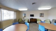This party room is a versatile, single-floor gathering space designed to accommodate a variety of social events and functions. The room features a comfortable lounge area furnished with cushioned green armchairs arranged around a circular coffee table, creating an inviting setting for relaxed conversation. A wall-mounted flat-screen television is positioned above a dark wood credenza, making the space well-suited for both casual viewing and presentations. Complementing the lounge area, round dining tables paired with blue chairs provide additional seating for guests, offering flexible arrangements to suit different event formats. Natural light filters into the room through a window dressed with neutral curtains, contributing to the room's bright and airy atmosphere. Recessed fluorescent lighting in the drop-tile ceiling ensures consistent, even illumination throughout. Colorful framed artwork decorates the walls, adding a touch of character to the neutral beige tones of the room. The carpeted flooring adds warmth and comfort underfoot, while the overall layout is open and functional, making this space an ideal venue for parties, community gatherings, and social events of all kinds.