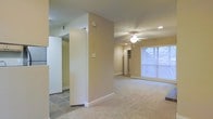 This single-floor space features an open, functional layout with a clear distinction between living and transitional areas. The main living area is finished with wall-to-wall carpet, lending a comfortable and quiet underfoot feel, while adjacent transitional spaces showcase tile flooring for easy maintenance. Warm neutral-toned walls run throughout, creating a cohesive and understated aesthetic.

A ceiling fan with an integrated light fixture is centrally positioned in the living area, complementing recessed lighting that provides even, ambient illumination across the space. A large window along the far wall allows generous natural light to fill the room, enhancing its open feel.

A structural column divides portions of the interior, subtly defining separate zones without fully enclosing them. Visible built-in shelving or closet storage can be seen toward the rear of the space, offering practical organization options. The textured ceiling adds subtle dimension overhead. Overall, the space presents a clean, move-in-ready environment with straightforward, practical finishes throughout.