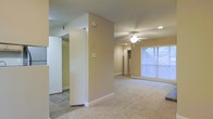 This single-floor space features an open, functional layout with a clear distinction between living and transitional areas. The main living area is finished with wall-to-wall carpet, lending a comfortable and quiet underfoot feel, while adjacent transitional spaces showcase tile flooring for easy maintenance. Warm neutral-toned walls run throughout, creating a cohesive and understated aesthetic.

A ceiling fan with an integrated light fixture is centrally positioned in the living area, complementing recessed lighting that provides even, ambient illumination across the space. A large window along the far wall allows generous natural light to fill the room, enhancing its open feel.

A structural column divides portions of the interior, subtly defining separate zones without fully enclosing them. Visible built-in shelving or closet storage can be seen toward the rear of the space, offering practical organization options. The textured ceiling adds subtle dimension overhead. Overall, the space presents a clean, move-in-ready environment with straightforward, practical finishes throughout.