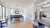 This single-floor entertainment lounge in Memphis, Tennessee, is designed with a contemporary aesthetic, combining open-concept layout with thoughtfully defined activity zones. The space is anchored by warm hardwood-style flooring that flows throughout, complemented by crisp white walls and high ceilings that enhance the sense of openness and airiness.

The recreational area features a full-size billiards table positioned beneath a striking black candelabra-style chandelier, surrounded by large black-framed windows that frame views of the surrounding neighborhood and flood the space with natural light. Bistro-style seating nearby provides a casual spot for socializing.

The kitchen is a standout feature, finished with gray cabinetry, open floating shelves styled with greenery and decorative accents, a tile backsplash, and a large island clad in navy hexagonal tile topped with a sleek gray countertop. Globe pendant lights illuminate the prep and serving area, while a stainless steel refrigerator and full sink add practical functionality.

A dedicated dining zone features a long upholstered banquette in rich burgundy, paired with dark bistro tables and chairs set against floor-to-ceiling windows with outdoor access. An adjacent lounge area is visible through a connecting doorway, completing this versatile communal space.