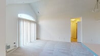 This single-floor residence offers a bright, open layout defined by clean lines and neutral tones throughout. The living room is a standout space, featuring soaring vaulted ceilings that amplify the sense of volume and a large arched window fitted with vertical blinds, allowing generous natural light to fill the room. Plush wall-to-wall carpeting runs through the living area, adding warmth underfoot. A peninsula counter connects the living space to the kitchen, which is outfitted with white cabinetry featuring sleek bar-pull hardware, light countertops, a coil-burner electric range, a refrigerator, and a dishwasher. The tile flooring in the kitchen provides a practical contrast to the carpeted living zones. The bedrooms continue the home's neutral palette, each carpeted and equipped with vertical-blind windows that bring in outdoor views and natural light. The bathroom features a full tub and shower combination with white square-tiled surround, chrome fixtures, a curved shower rod, mosaic tile flooring, and a toilet and vanity in a clean, well-maintained layout. Overall, the space presents a bright, functional, and move-in-ready environment with a cohesive design sensibility.