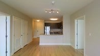 This single-floor space showcases a contemporary open-concept design that seamlessly connects the living area to the kitchen. The kitchen is a standout feature, equipped with stainless steel appliances, including a full-size refrigerator and built-in microwave. Dark espresso cabinetry provides a striking contrast against the light granite countertops, which extend onto a peninsula that doubles as a breakfast bar. This peninsula serves as both a functional workspace and a natural divider between the kitchen and the main living area.

The flooring throughout the space consists of light-toned hardwood-style planks, lending warmth and continuity to the open layout. The neutral beige walls complement the overall modern aesthetic. Lighting is well-considered, with a multi-bulb track light fixture in the main living area and flush-mount ceiling lights providing even illumination throughout. The space features multiple doorways leading to additional rooms, suggesting a practical and efficient floor plan. Clean white baseboards and a smooth ceiling finish add to the polished, move-in-ready feel of this well-designed unit.