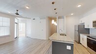 This well-designed single-floor residence offers a clean, modern aesthetic throughout. The heart of the home is an open-concept kitchen and living area, where a striking island with a teal base and granite countertop takes center stage. Pendant lights in brushed gold hang above, casting warm, focused light over the space. The kitchen is fully equipped with stainless steel appliances — including a refrigerator, electric range, and built-in microwave — framed by tall white cabinetry with sleek hardware and a classic subway tile backsplash. A built-in shelving and desk nook adds a functional touch nearby. Light wood-style flooring flows throughout the main living areas, enhancing the bright, airy atmosphere created by white walls and recessed lighting. The bedrooms are carpeted for comfort, each featuring a ceiling fan and windows with blinds that invite natural light and outdoor views. The bathroom is thoughtfully appointed with a granite-topped vanity, white cabinetry, a large mirror, and a tub-shower combination with white subway tile surround. A walk-in closet is accessible directly from the bathroom, adding practical storage. Overall, the space presents a cohesive and contemporary design with quality finishes throughout.