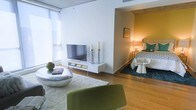 This single-floor space showcases a thoughtfully designed open-concept layout that seamlessly connects the living and sleeping areas. The living space is anchored by hardwood flooring and features large windows fitted with roller shades, allowing abundant natural light to brighten the interior. A wall-mounted media console with a flat-screen television serves as the focal point, complemented by decorative candle holders and curated accessories. A plush white area rug defines the seating zone, where a distinctive green curved chair and a rounded metallic coffee table add modern character to the space.

A partial dividing wall creates a semi-private sleeping alcove, bathed in warm ambient lighting from a bedside lamp. The sleeping area features a neatly styled bed with teal accent pillows, a teal area rug, and a small side table. The accent wall in the sleeping nook is painted in a rich mustard yellow, adding a warm contrast to the otherwise neutral palette throughout the space. The combination of natural light, warm artificial lighting, and contemporary furnishings creates an inviting and cohesive ambiance.