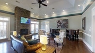 This single-floor clubhouse is thoughtfully designed to provide a comfortable and inviting gathering space for residents and guests. The main lounge area is anchored by a striking floor-to-ceiling stone fireplace, above which a large flat-screen television is mounted, making it an ideal spot for both relaxed evenings and communal viewing. Recessed lighting and a ceiling fan overhead ensure the space remains well-lit and comfortable year-round. Rich hardwood flooring runs throughout, adding warmth and a classic aesthetic to the interior. The seating area features a plush leather sofa paired with patterned accent chairs and a rustic wooden coffee table, creating an approachable and eclectic style. A decorative area rug grounds the lounge arrangement, while contemporary artwork adorns the walls. Toward the back of the space, a high-top bar seating area with dark wood chairs provides an alternative setting for casual dining or conversation. Tall French doors along the rear wall flood the interior with natural light and offer access to an outdoor patio area, seamlessly connecting the indoor and outdoor environments. A tasteful green accent border runs along the upper walls, adding a distinctive architectural detail that ties the room's color palette together. Overall, the clubhouse presents a polished yet relaxed ambiance suited for everyday use.