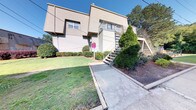 This residential building presents a classic angular architectural design characterized by its multi-level structure and neutral beige exterior. The facade features a combination of siding and stucco-style finishes, giving the building a sturdy, timeless appearance. An exterior metal staircase leads to an upper-level deck or balcony, which is fitted with a railing and offers an elevated outdoor area accessible from the upper floor. Windows are positioned throughout the exterior, allowing for natural light to enter the interior spaces.

The grounds surrounding the building are thoughtfully landscaped, with a neatly trimmed lawn, decorative shrubs, and mature evergreen trees that provide natural privacy and greenery. A concrete walkway extends from the building's entrance, providing clear and direct pedestrian access to and from the property. The landscaping includes well-maintained hedgerows along the building's base, adding to the overall curb appeal.

The setting is complemented by a backdrop of tall, mature trees that frame the property and contribute to a serene, established neighborhood atmosphere. The outdoor areas, combined with the building's clean architectural lines, create a functional and visually cohesive exterior environment.