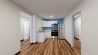 This efficiently designed single-floor unit offers a thoughtful layout across its kitchen, hallway, bathroom, and living area. Rich, wood-look vinyl plank flooring flows consistently throughout the space, lending warmth and visual continuity to every room. The kitchen is a standout feature, showcasing white shaker-style cabinetry, granite-look countertops, and a full suite of stainless steel appliances including a range, over-the-range microwave, and refrigerator. A vibrant blue accent wall adds a bold pop of color, complementing the clean white cabinetry. The living area is bright and open, featuring a large window with a roller shade that allows natural light to fill the room, along with a ceiling fan for added comfort. A wall-mounted PTAC unit provides climate control. The hallway connects the spaces efficiently, with a blue-trimmed entry door adding a subtle design accent. The bathroom is clean and functional, featuring light tile flooring, a floating wood-toned vanity with a white countertop and undermount sink, a framed mirror, and chrome fixtures. The overall aesthetic is modern and clean, with a neutral white palette punctuated by thoughtful color accents.