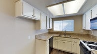 This single-floor residence offers a well-organized and functional layout designed for comfortable everyday living. The kitchen is a standout feature, showcasing white cabinetry that extends to the upper walls, providing generous storage throughout. Granite-style countertops offer durable workspace, while a double-basin sink sits beneath a window fitted with horizontal blinds, allowing natural light to filter in. A built-in dishwasher adds modern convenience to the space. Overhead, a large skylight-style flush light fixture brightens the kitchen with ample illumination, complementing the natural light from the window. The rich, dark wood-look flooring grounds the space with warmth and contrast against the lighter cabinetry and walls. The neutral beige wall tones throughout contribute to a clean, cohesive aesthetic. The textured ceiling adds a classic residential character to the interior. Overall, the space presents a practical and well-maintained environment with thoughtful design details, making it well-suited for those seeking a move-in-ready home with functional amenities and a timeless aesthetic.