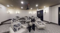 Located in Huntersville, NC, this single-floor screening room is thoughtfully designed to deliver a premium private cinema experience. The main theater area is anchored by a large framed projection screen mounted on a neutral-toned accent wall, paired with a ceiling-mounted projector positioned at the center of the room. Recessed lighting and sleek wall sconces cast a warm, ambient glow throughout the space, complementing the dark ceiling that enhances the cinematic atmosphere. The seating arrangement features multiple plush, light-upholstered armchairs adorned with decorative accent pillows, organized around dark-finished side tables — providing a comfortable and stylish viewing environment for groups. Soft, patterned carpet in neutral grey tones extends throughout, adding warmth underfoot and contributing to the room's acoustic quality. A foyer-style entry corridor connects to the main screening area and includes a small refreshment station equipped with a beverage dispenser and a stainless steel waste bin, offering added convenience for guests. Dark-trimmed doors and clean, modern baseboards throughout the space contribute to its cohesive, contemporary aesthetic. Large windows along one wall allow for views of the surrounding exterior while maintaining the room's refined, upscale character.