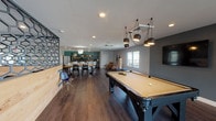 The Halsey Flats community amenity space is a thoughtfully designed, single-floor gathering area that seamlessly blends recreation, dining, and relaxation into one open, flowing layout. Dark hardwood-style flooring runs throughout, providing a cohesive foundation that ties the various zones together.

The game room anchors one end of the space, centered around a full-size pool table beneath a striking cluster pendant light fixture. A wall-mounted flat-screen television and a dedicated cue rack add functional detail, while an accent wall in charcoal grey lends a modern edge. Adjacent to the game room, a comfortable lounge area features a textured dark accent wall, warm seating in muted tones, and recessed lighting that creates an inviting atmosphere.

The dining zone offers a substantial live-edge wood communal table surrounded by black chairs, positioned beneath a simple pendant light. Behind it, a full kitchen outfitted with stainless steel appliances, a teal accent wall, open shelving, and a breakfast bar provides a practical space for entertaining and shared meals.

At the far end of the space, large sliding glass doors open to an outdoor deck, flooding the interior with natural light and offering open views of the surrounding neighborhood. This bright transitional area features additional lounge seating and creates a welcoming connection between indoors and outdoors.